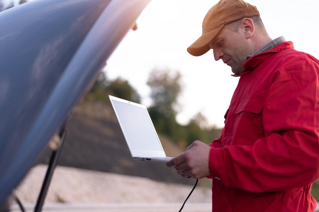 Car hood open with man in ballcap and red coat holding a laptop computer connected to car engine computer